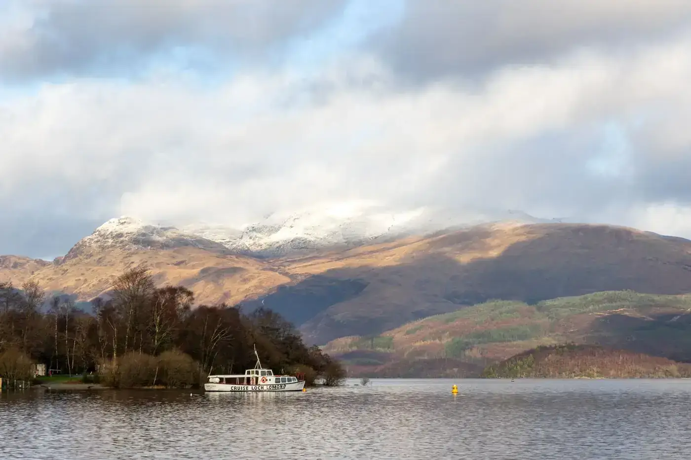 A scenic winter view of Loch Lomond with a boat and snow-capped mountains in Scotland.