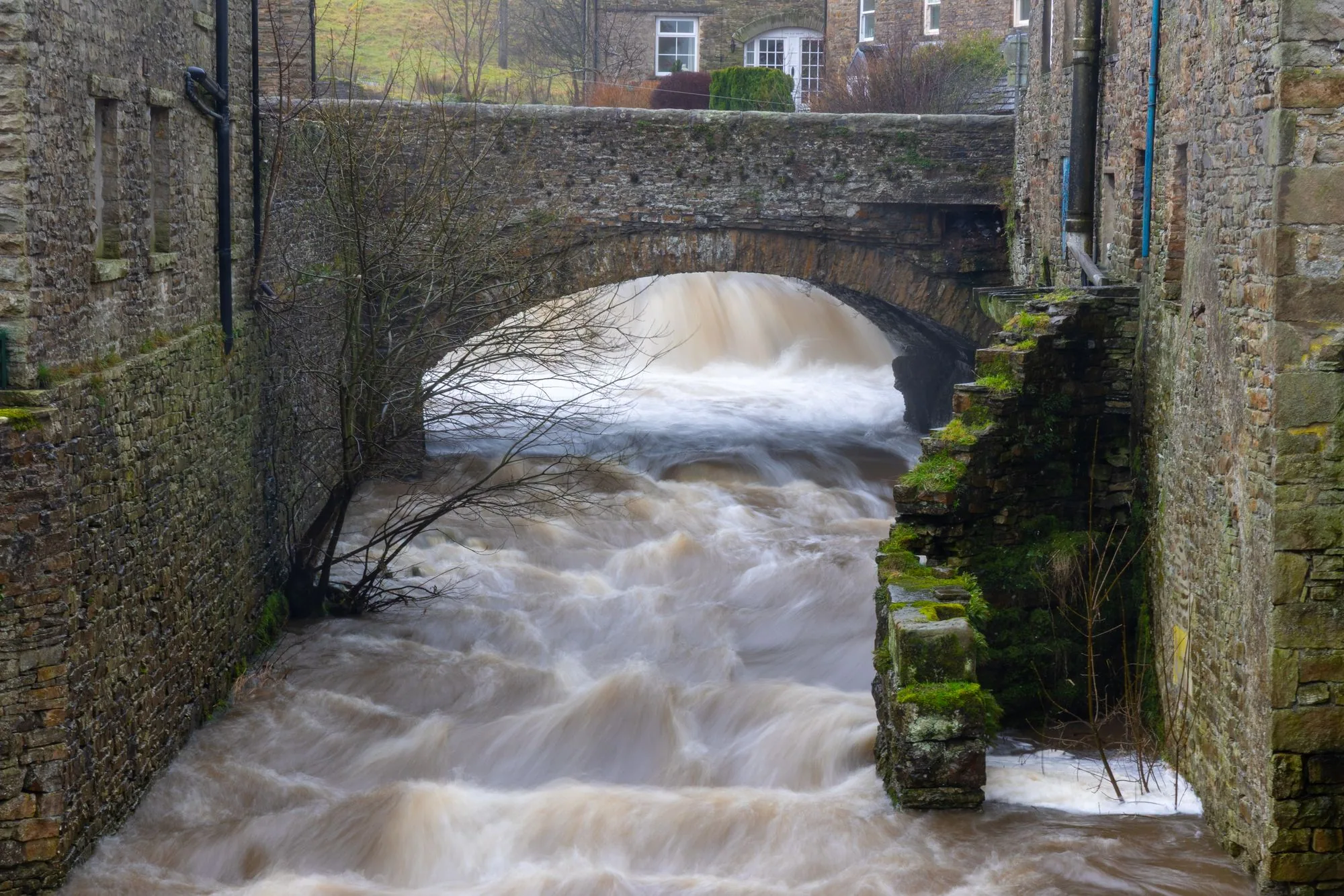 Brown water rushes under a historic stone bridge in Hawes, UK, showcasing winter's powerful flow.