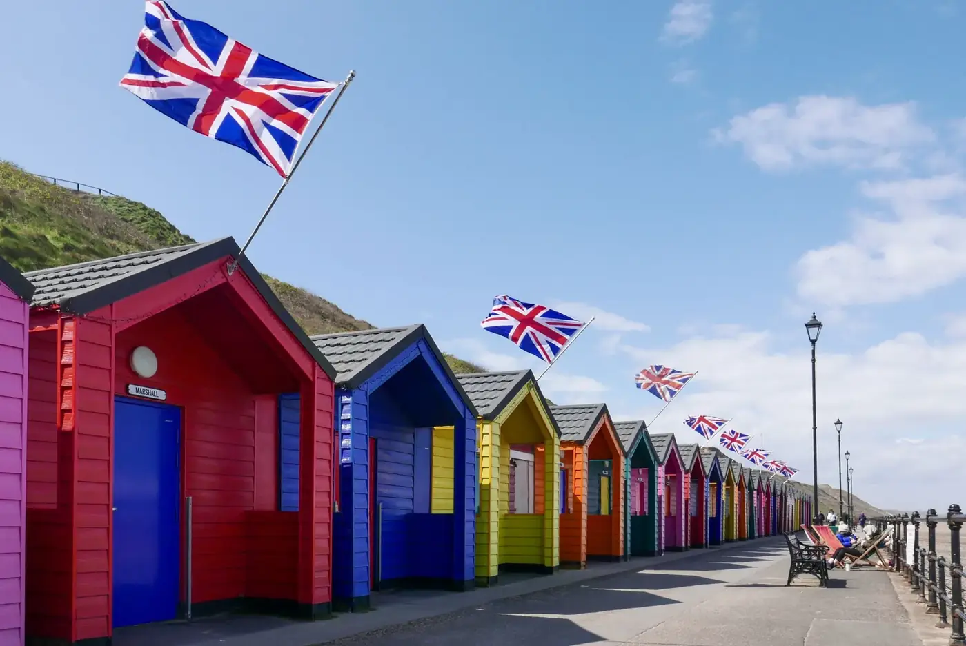Colorful beach huts with Union Jack flags in Saltburn-by-the-Sea, England, under a clear blue sky.